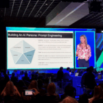 A conference with an image of a slideshow and a woman speaking projected onto a large screen.