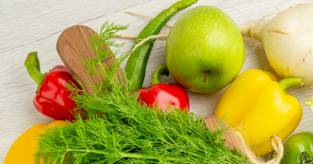 Fresh vegetables and fruit including bell peppers, green apple, onion, chili peppers, and dill on a wooden surface.