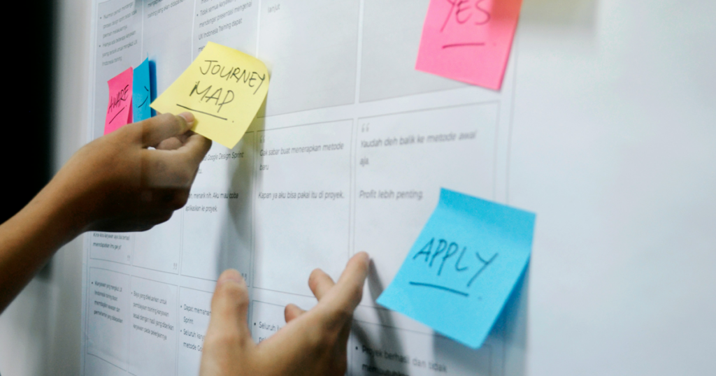 A person placing sticky notes labeled “Journey Map” and “Apply” on a planning board.