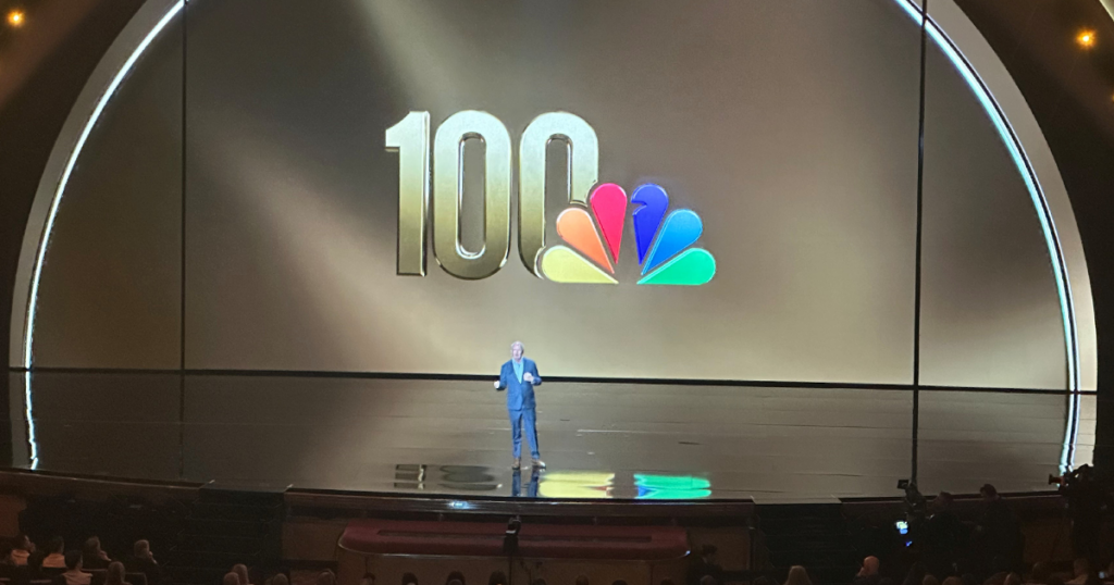 A presenter stands on a large stage in front of a giant screen displaying a “100” logo alongside the colorful NBC peacock logo.